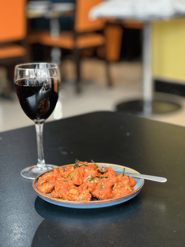 A blue plate of spicy fried chicken garnished with curry leaves, served next to a glass of red wine on a dark table