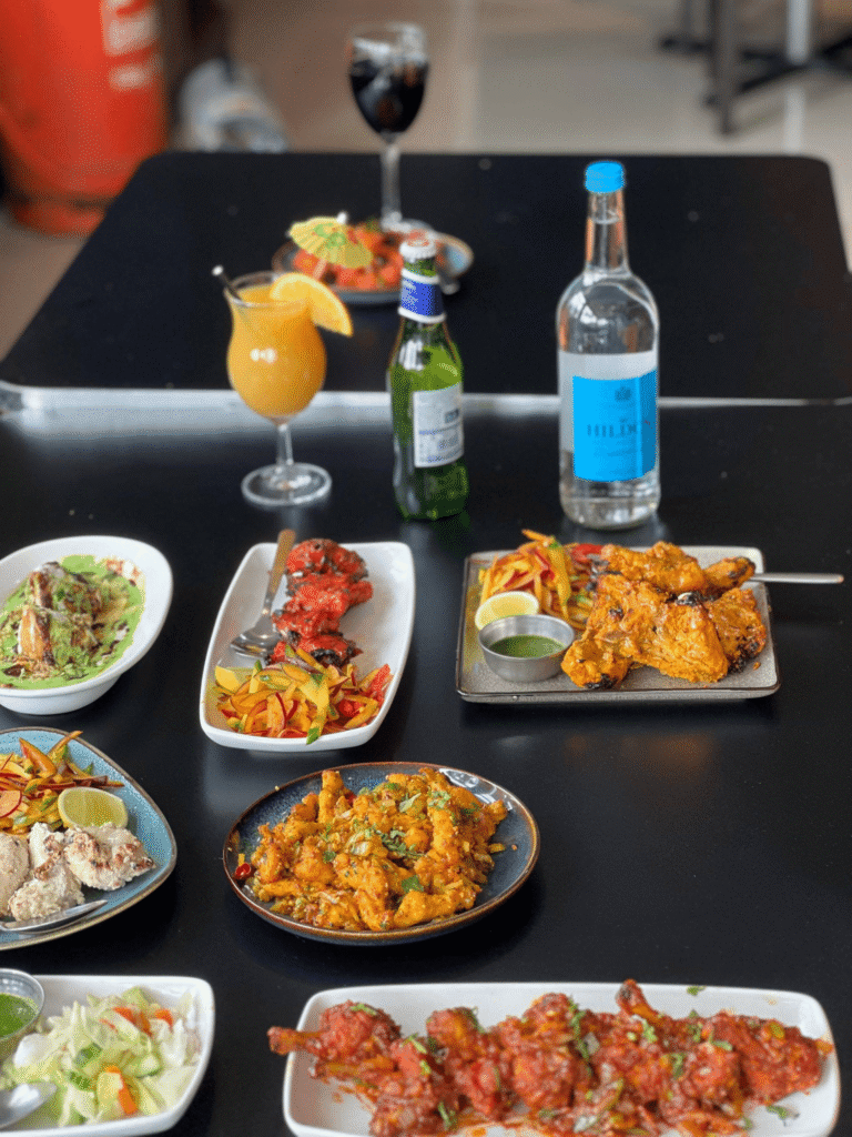 A dining table filled with a colorful selection of Indian dishes including chicken tikka, stir-fried starters, side salads, and various drinks