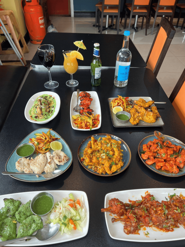 A dining table filled with a large feast of Indian dishes and drinks, including chicken tikka, spicy chicken wings, chaat, and a cocktail