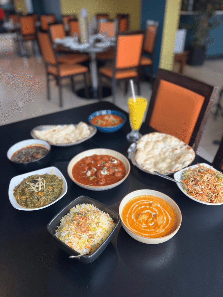 A dining table filled with an Indian dinner spread including butter chicken, palak paneer, biryani rice, naan bread, noodles, and a mango lassi