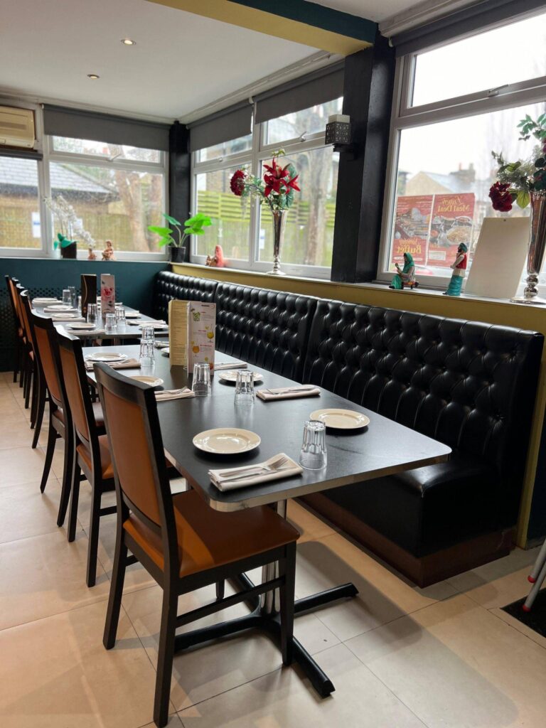 A modern restaurant dining area with set tables, orange chairs, and a long tufted black leather booth running alongside large windows