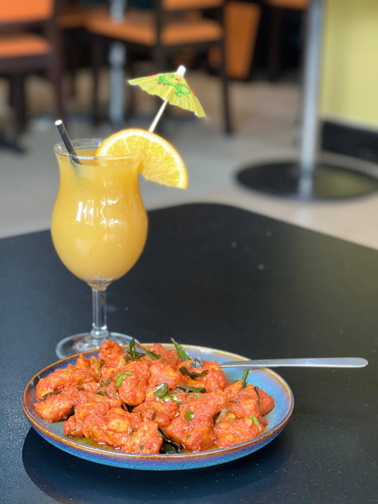 A plate of fried spicy red chicken garnished with herbs, served beside a yellow tropical cocktail topped with an orange slice