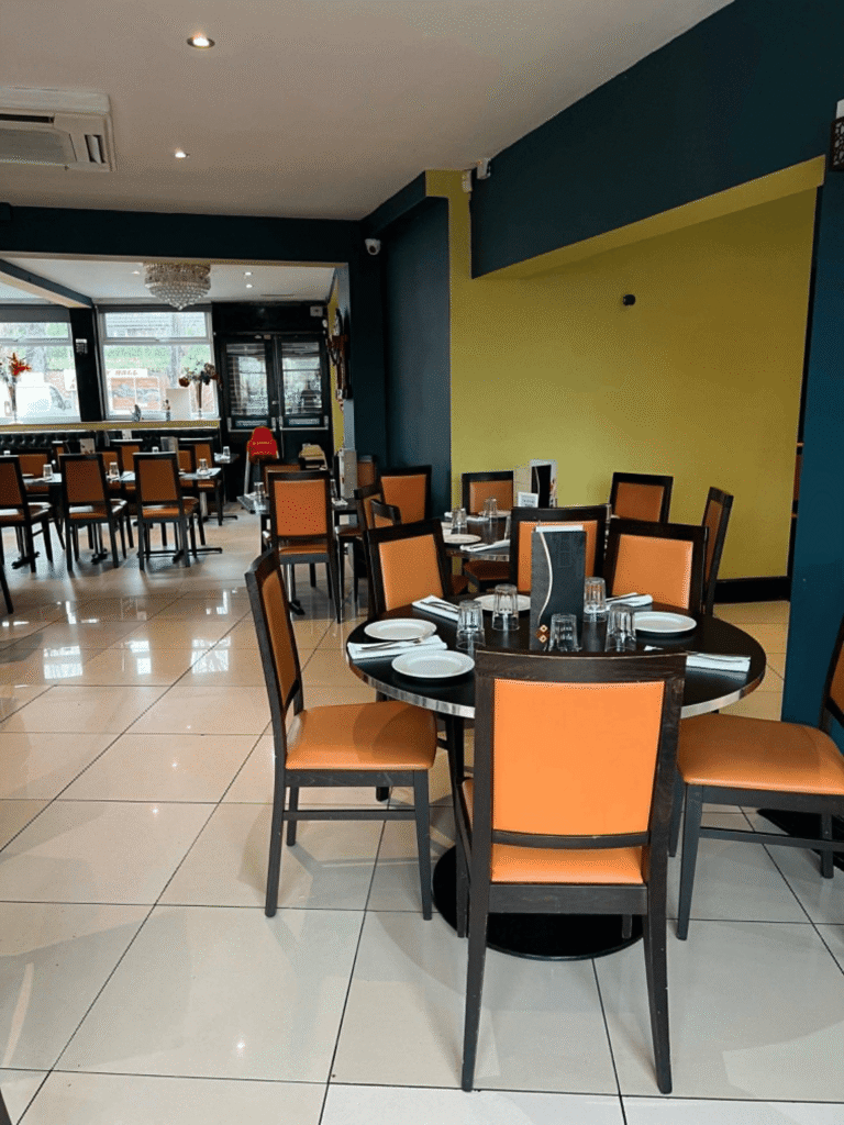 A spacious restaurant dining room featuring tiled floors, teal and mustard walls, and round dark wood tables surrounded by orange-upholstered chairs