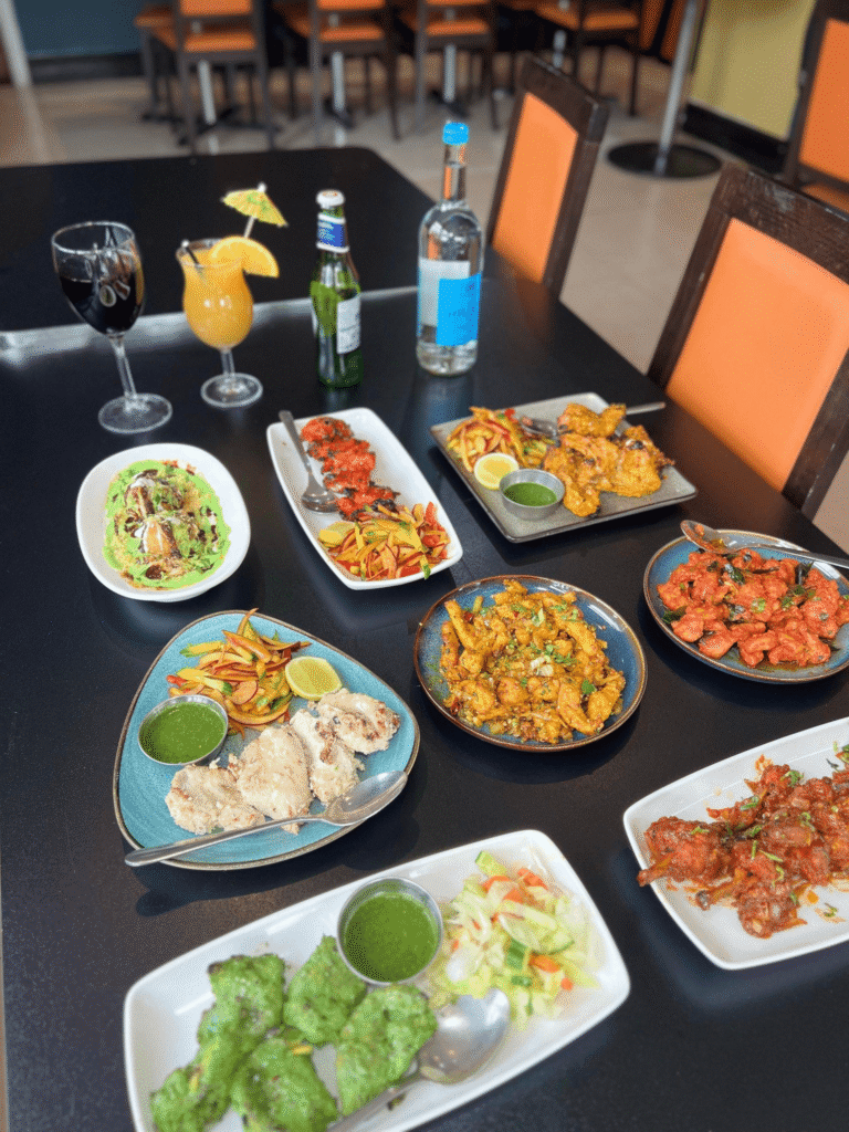 An angled view of a dining table filled with a large selection of Indian appetizers, grilled chicken tikka, spicy wings, chaat, and drinks