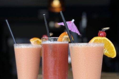 Three colorful cocktails arranged in a row with black straws, orange slices, a cherry skewer, and a pink paper umbrella